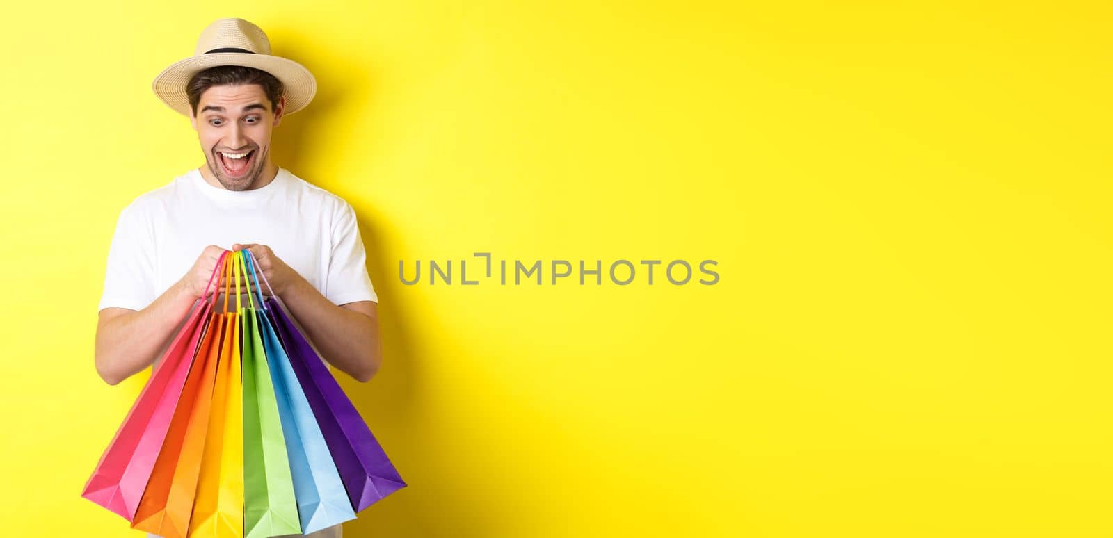 Image of happy man shopping on vacation, holding paper bags and smiling, standing against yellow background.