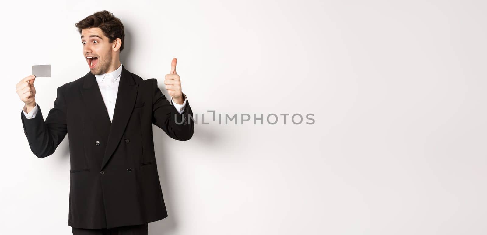 Image of excited handsome businessman, showing credit card and thumb-up, standing against white background by Benzoix