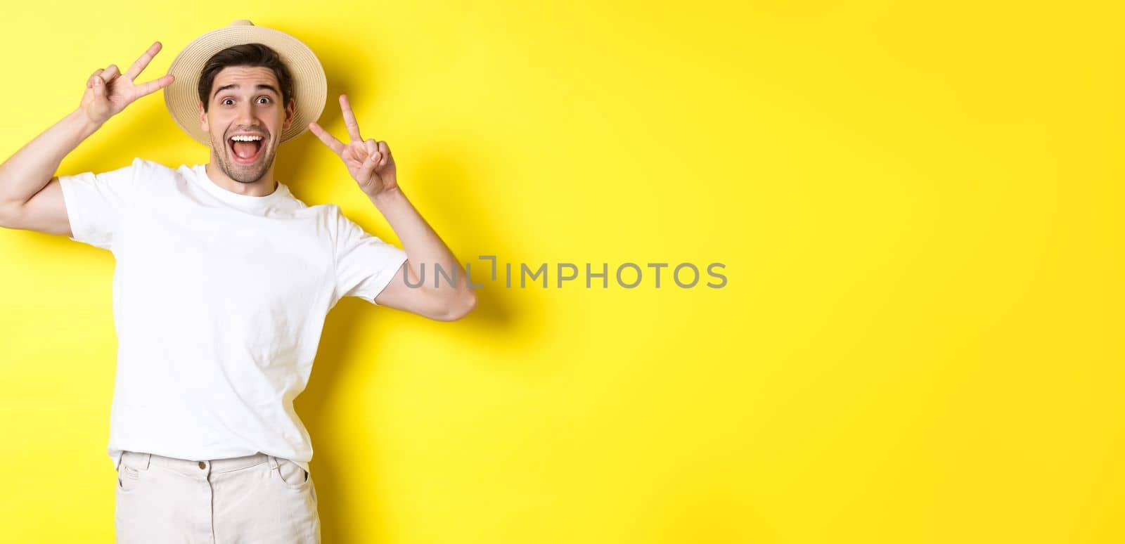 Concept of tourism and vacation. Happy male tourist posing for photo with peace signs, smiling excited, standing against yellow background.