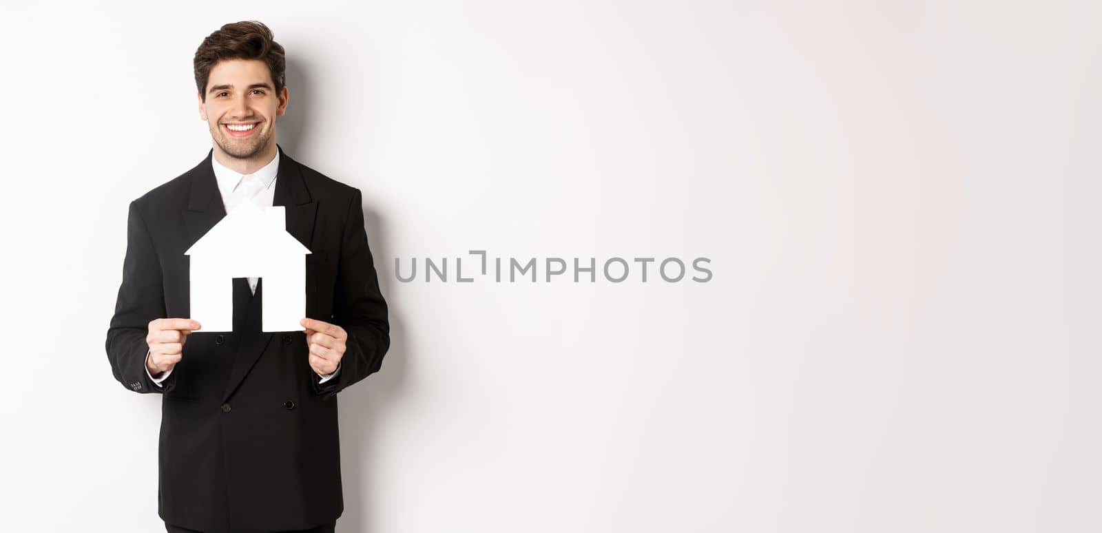 Image of handsome real estate agent in black suit showing home maket, smiling at camera, standing against white background.