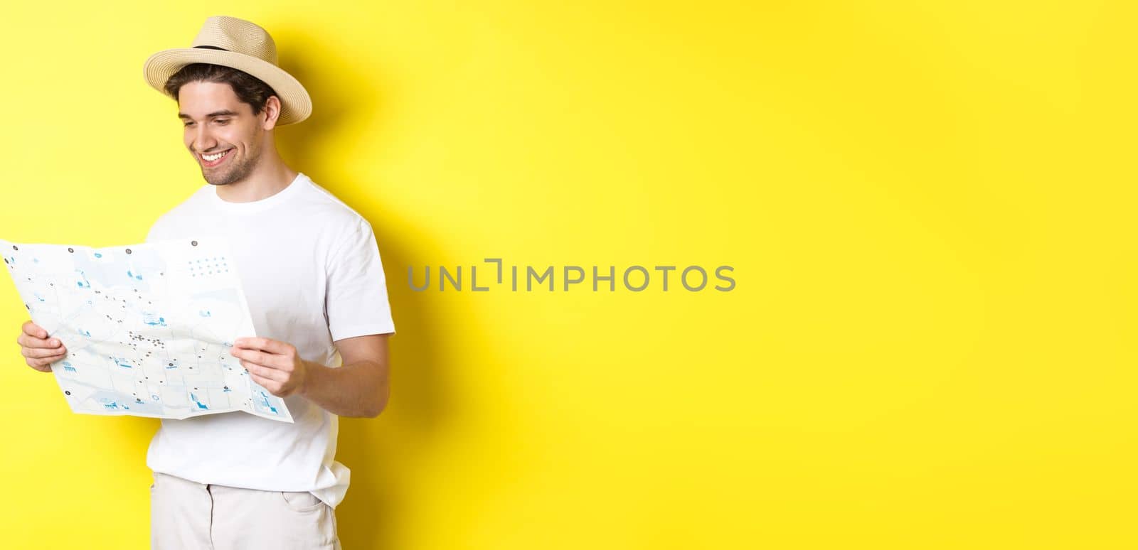 Travelling, vacation and tourism concept. Smiling happy tourist looking at map with sightseeings, standing against yellow background.