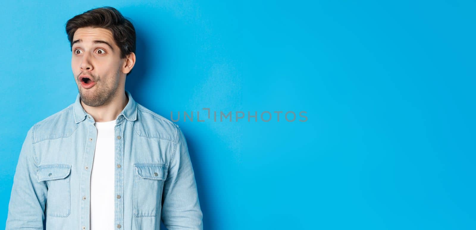 Close-up of surprised handsome man looking impressed left, open mouth fascinated, wearing casual outfit, standing over blue background.