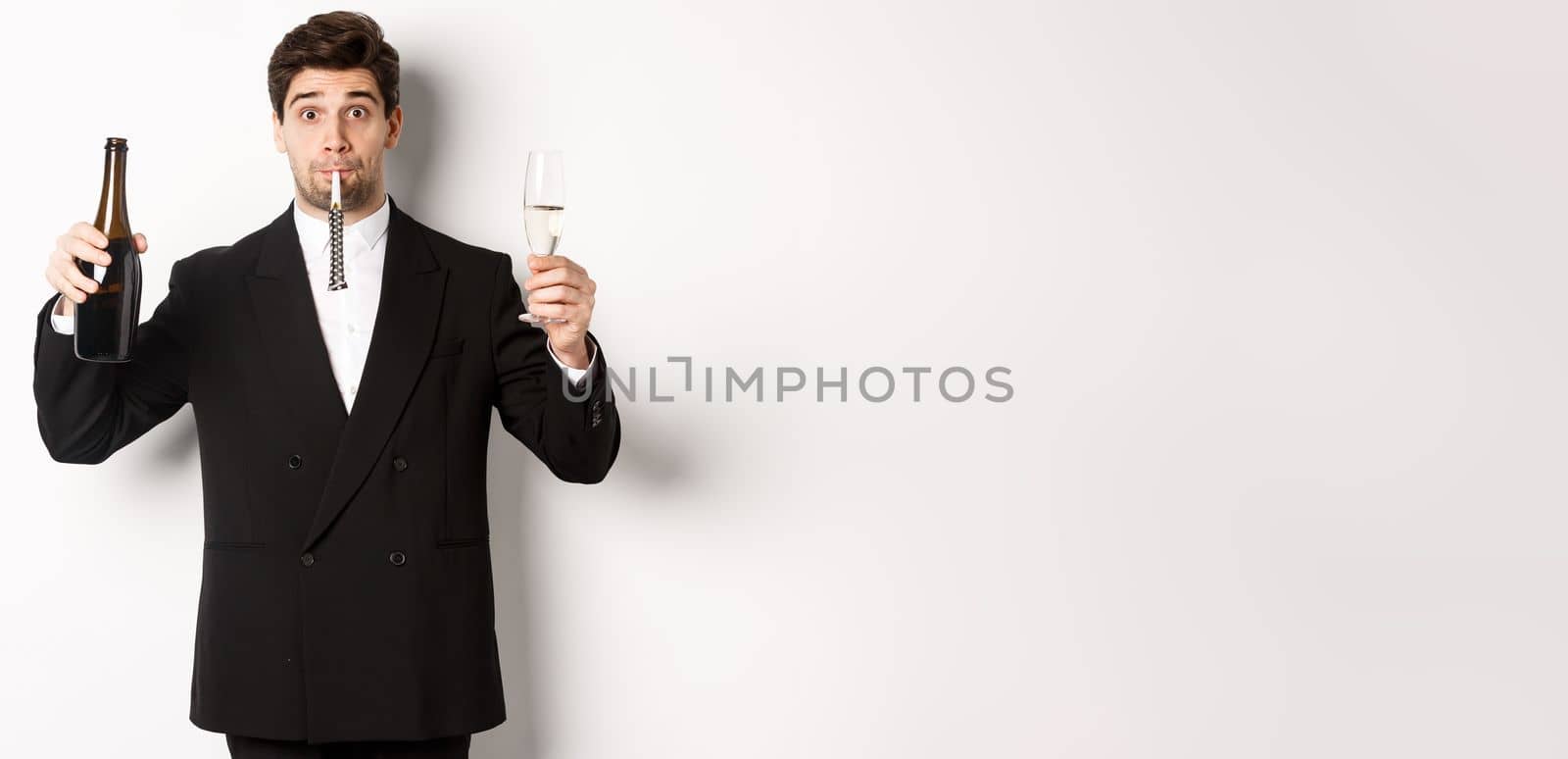 Concept of holidays, party and celebration. Portrait of handsome guy in black suit, raising bottle of champagne and glass, blowing a party whistle, having a birthday, standing over white background.