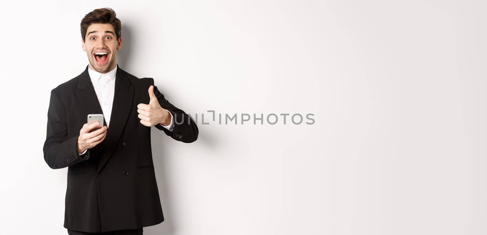 Portrait of handsome man in trendy suit showing thumbs-up in approval, using mobile phone app, smiling pleased, standing over white background.