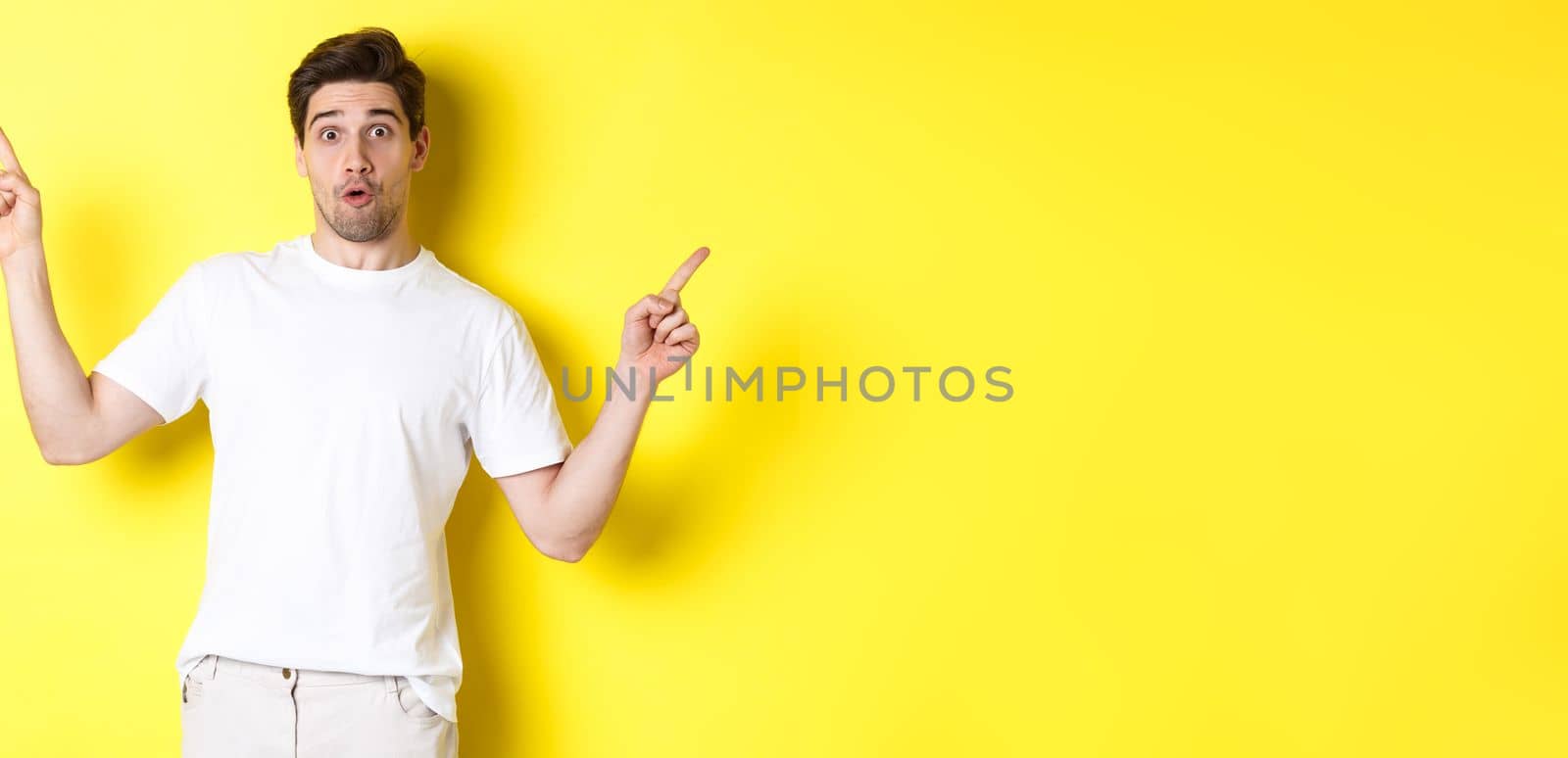 Handsome man pointing fingers sideways, showing two promos, standing over yellow background.