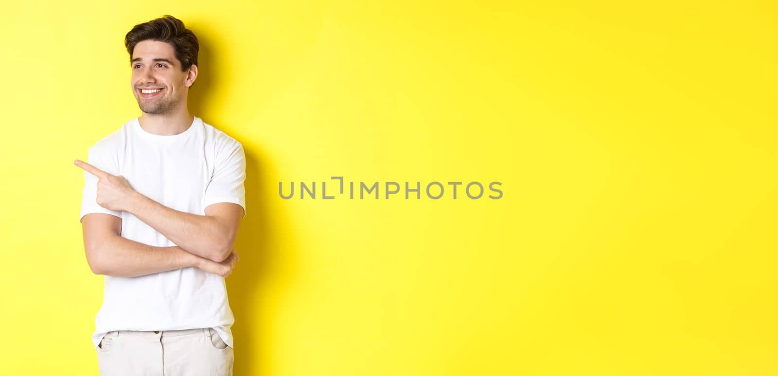 Handsome smiling man in white clothes, looking and pointing finger left at banner, standing over yellow background.