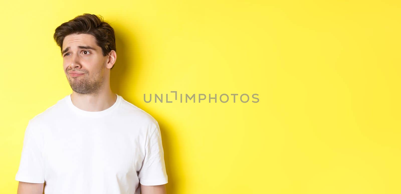 Reluctant guy in white t-shirt looking left, grimacing skeptical and displeased, standing over yellow background.