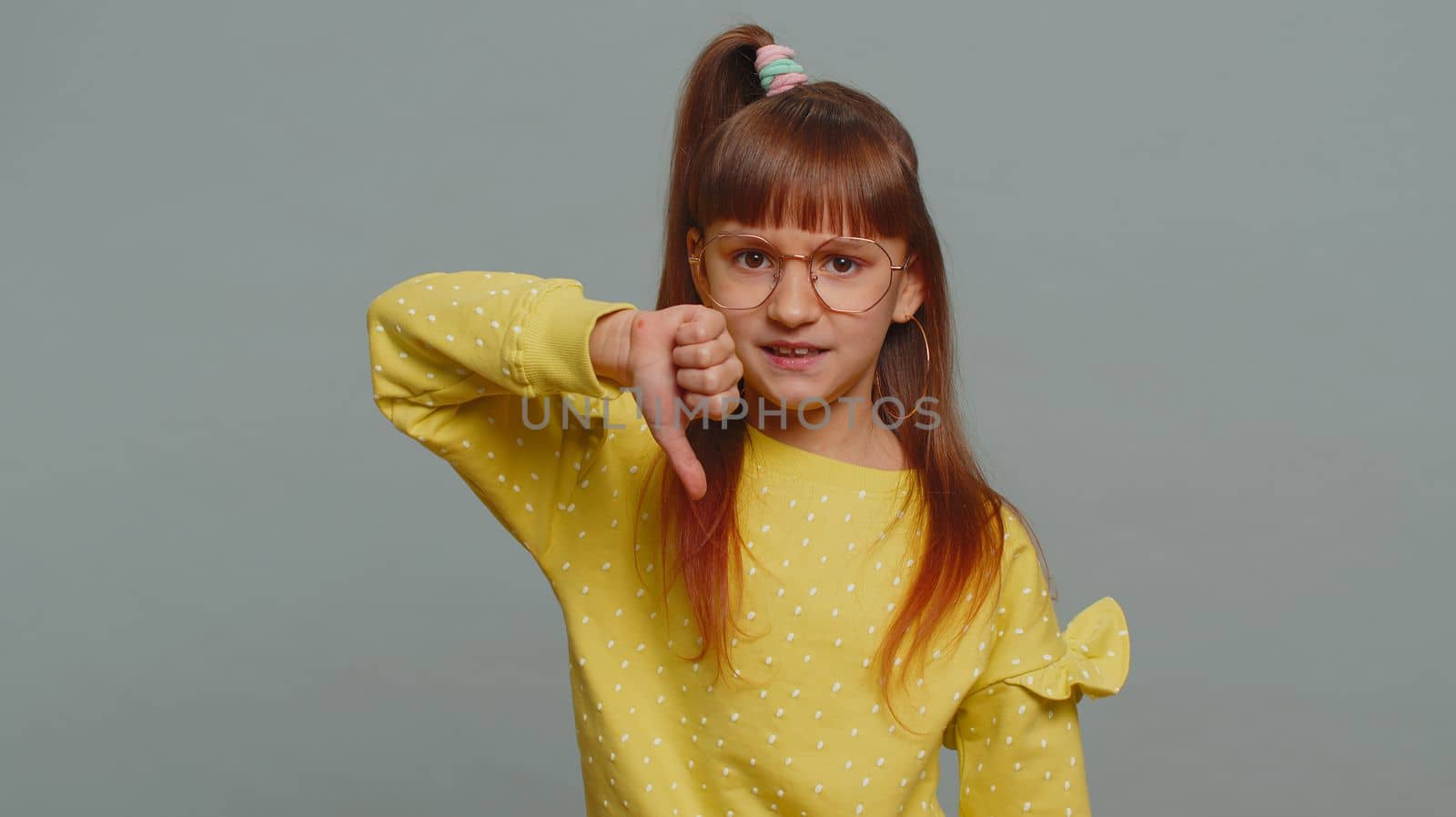 Dislike. Upset young preteen child girl kid showing thumbs down sign gesture, expressing discontent, disapproval, dissatisfied bad work indoors. Displeased toddler children on gray studio background