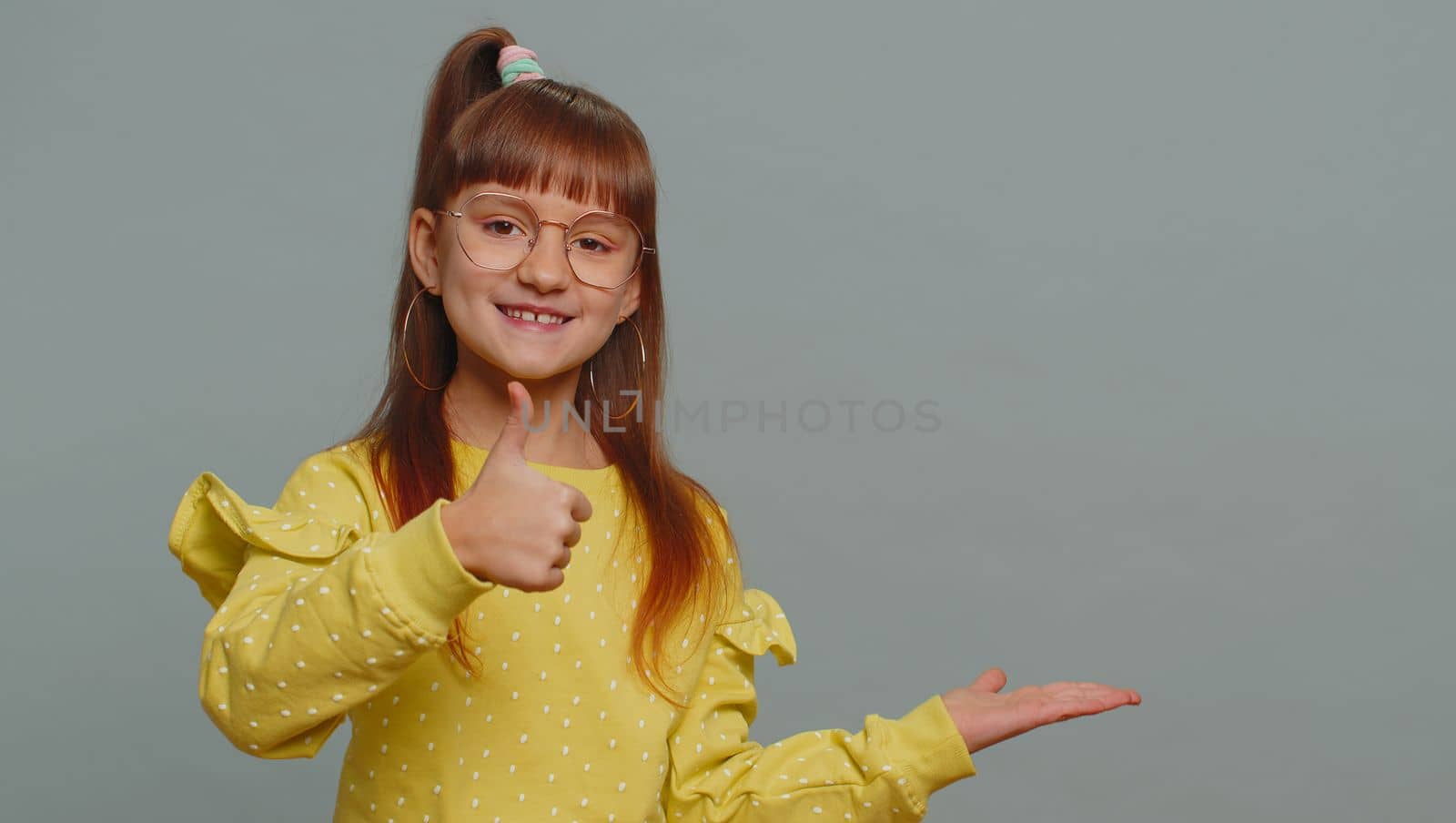 Happy toddler girl showing thumbs up pointing empty place, advertising area for commercial text, copy space for goods promotion banner, copy-space. Young child kid. Studio shot on gray wall background