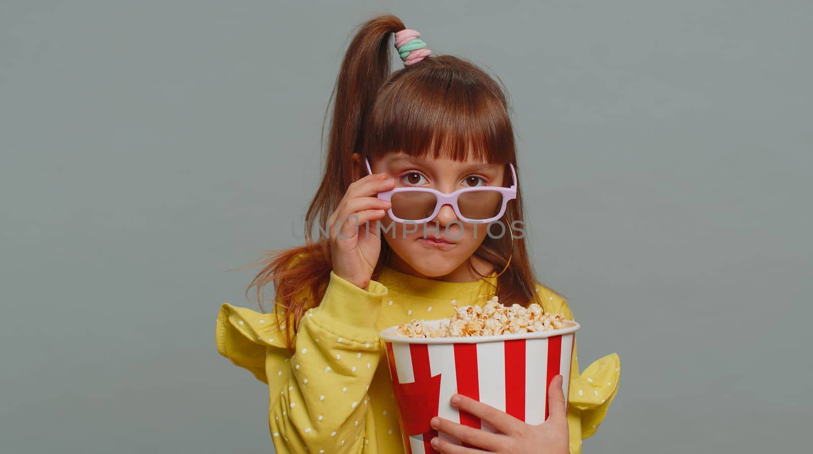 Excited young toddler school girl in 3D glasses eating popcorn, watching interesting tv serial, sport game, film, online social media movie content. Teen female child kid on studio gray background