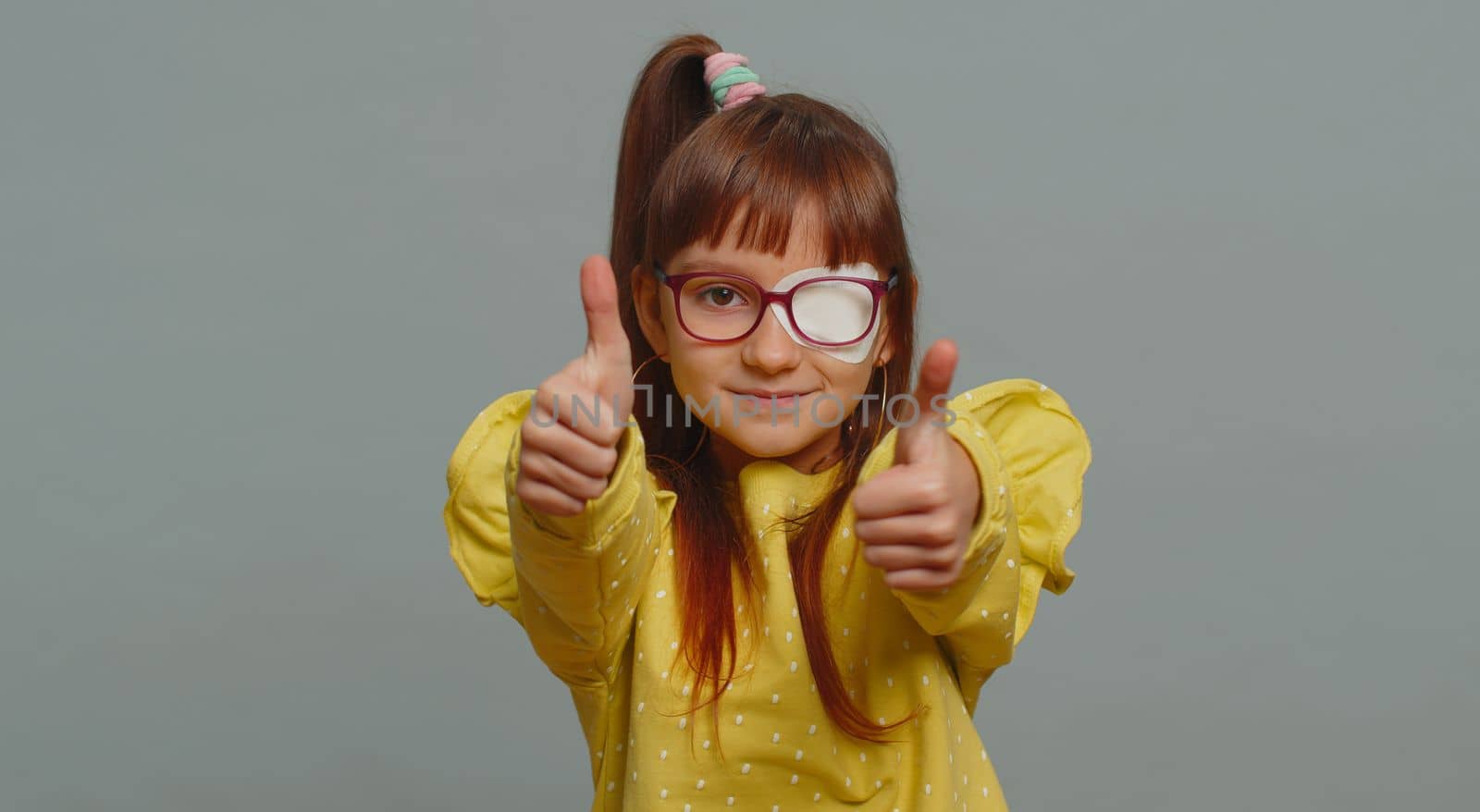 Like. Happy child girl kid having protective bandage plaster patch on one eye showing thumbs up like sign positive something good positive feedback. Toddler children isolated on studio gray background