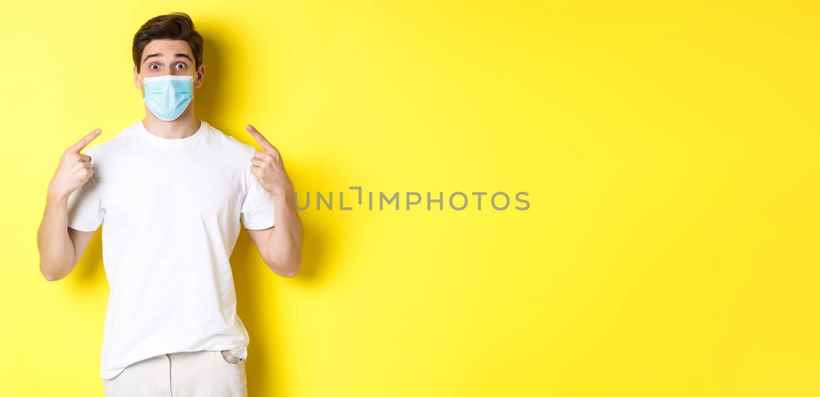 Concept of coronavirus, pandemic and social distancing. Young surprised man pointing at medical mask on face, yellow background. Copy space