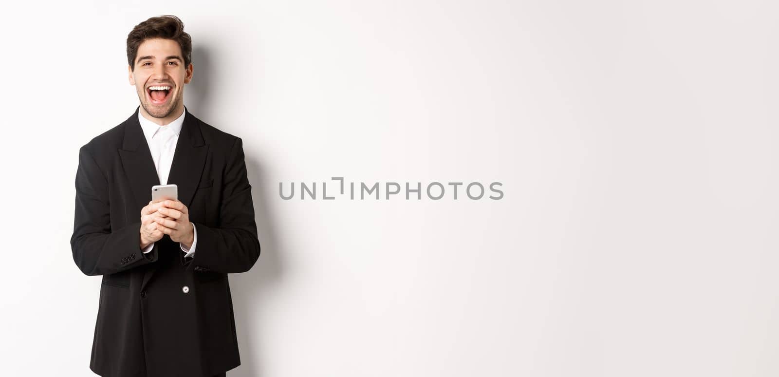 Portrait of happy good-looking man, wearing black suit, laughing from happiness and using mobile phone, standing over white background by Benzoix