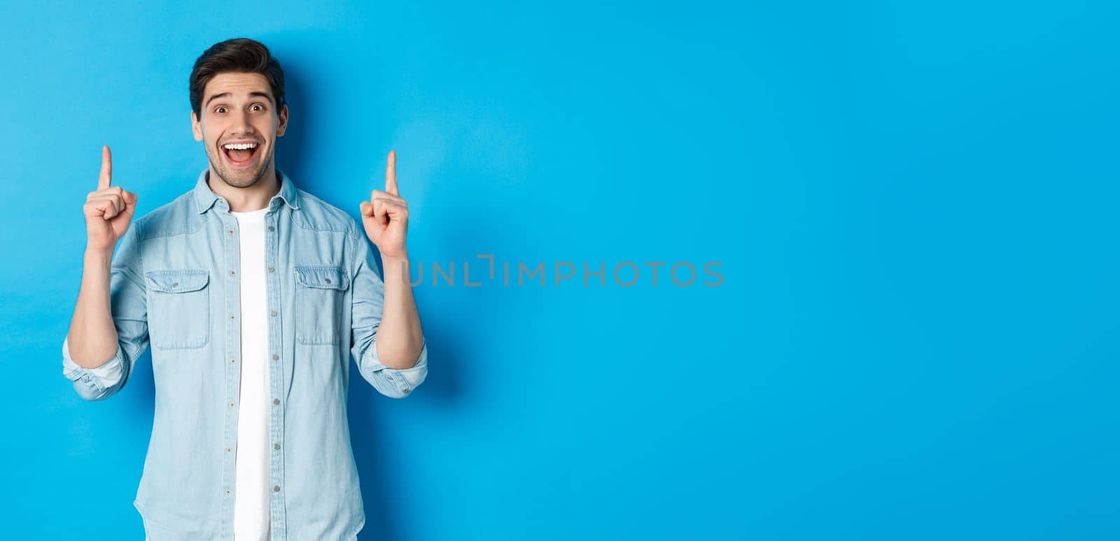 Portrait of happy 25s guy with beard, pointing fingers up and smiling, showing advertisement, standing against blue background.