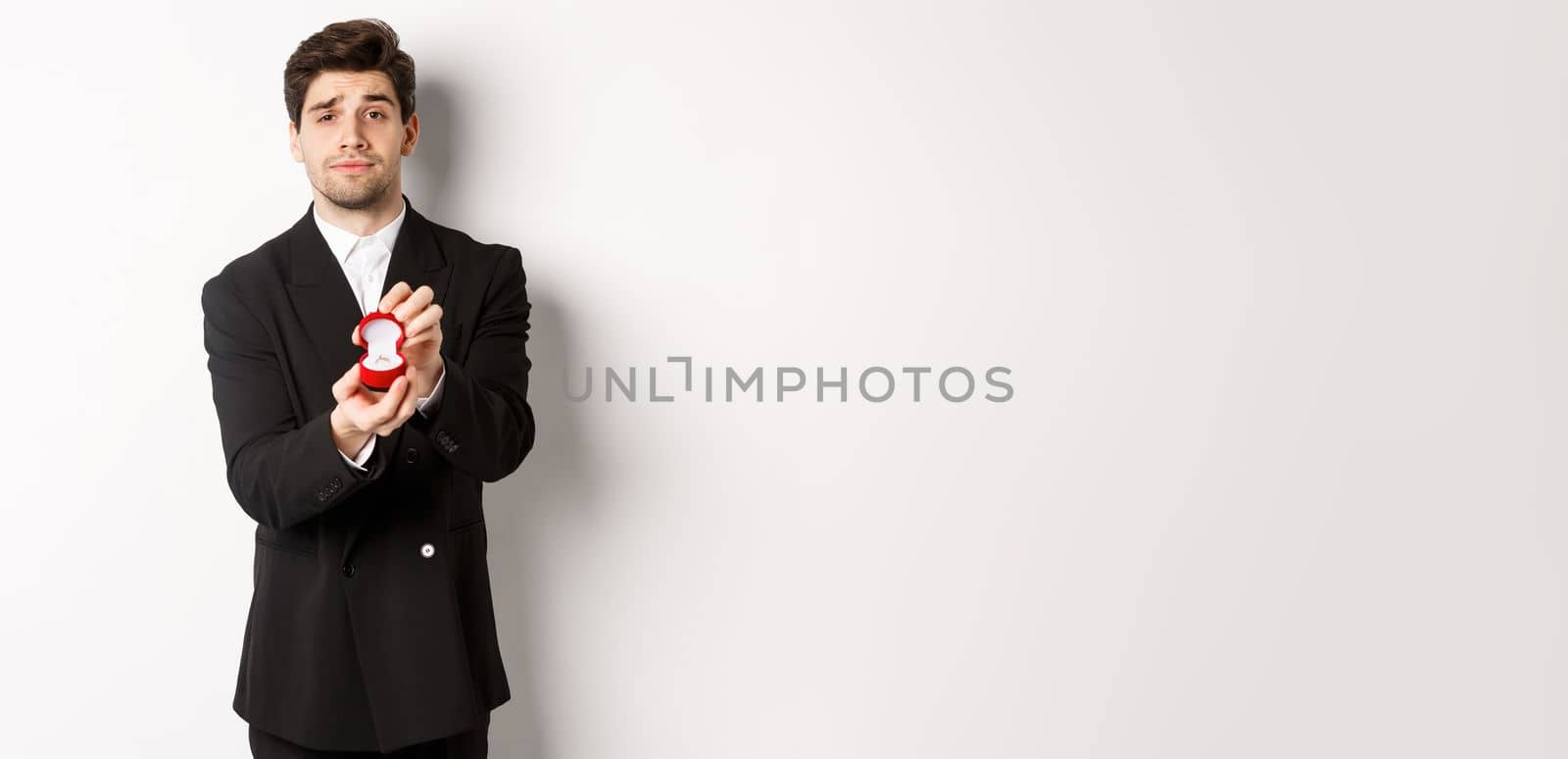 Portrait of handsome man making a proposal, asking to marry him, showing ring, standing in black suit against white background by Benzoix