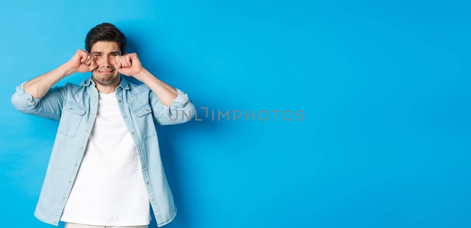 Sad timid man crying, wiping tears off face and sobbing, looking offended and upset, standing over blue background by Benzoix