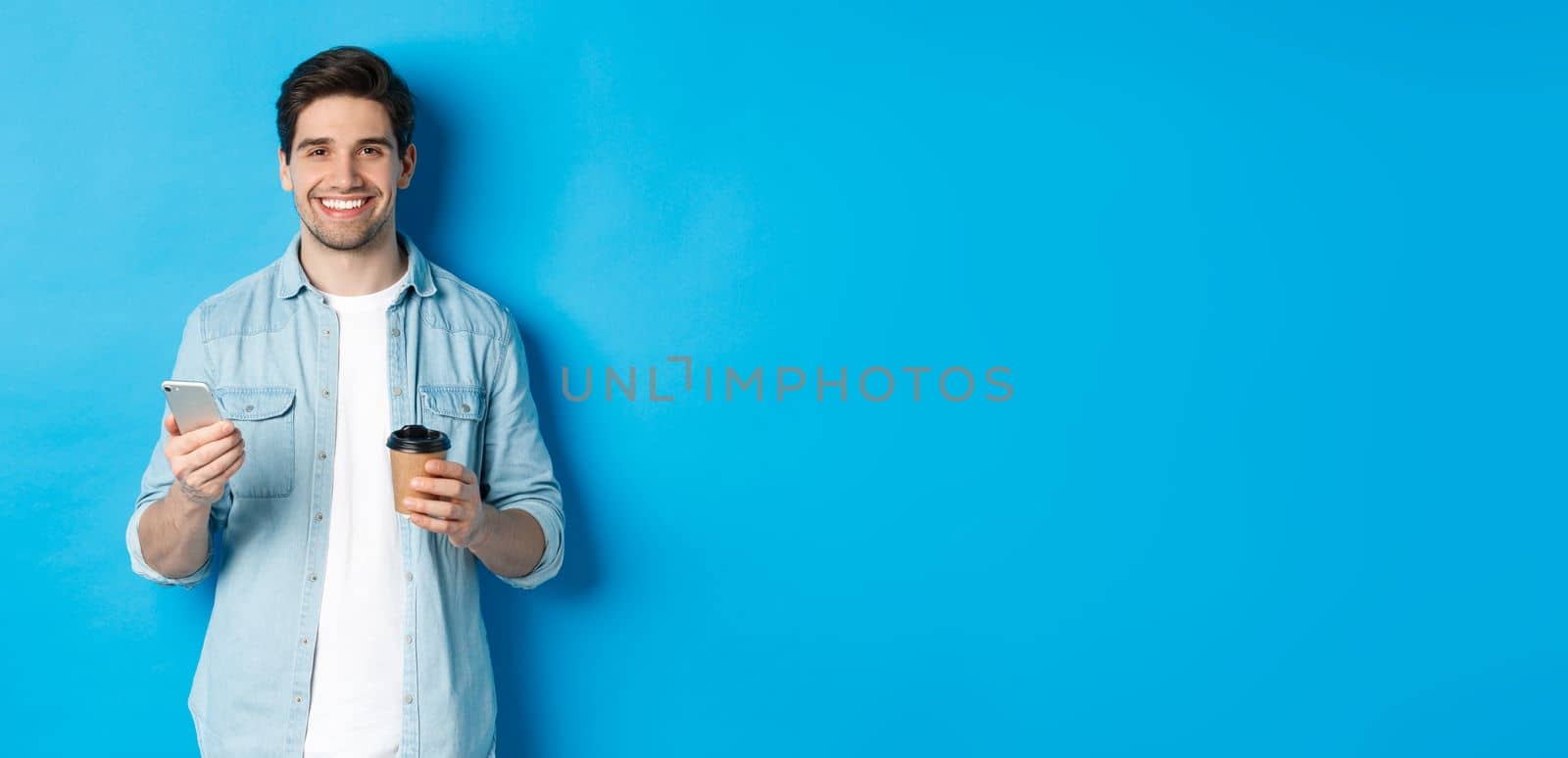 Young modern man drinking coffee and using mobile phone, smiling at camera, standing over blue background.