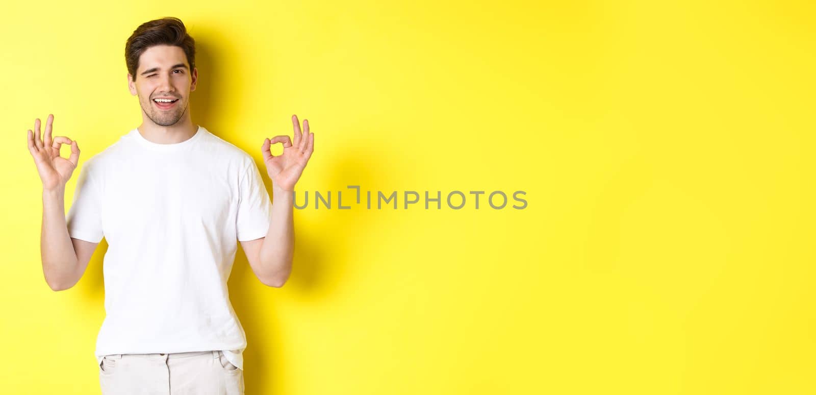 Confident handsome man winking, showing okay signs in approval, like something good, standing over yellow background.