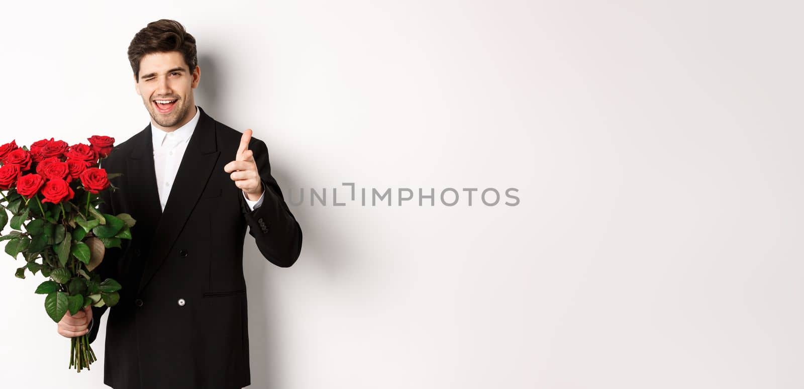 Image of handsome romantic guy in black suit, holding bouquet of roses and pointing at camera, congratulating with holiday, standing against white background by Benzoix