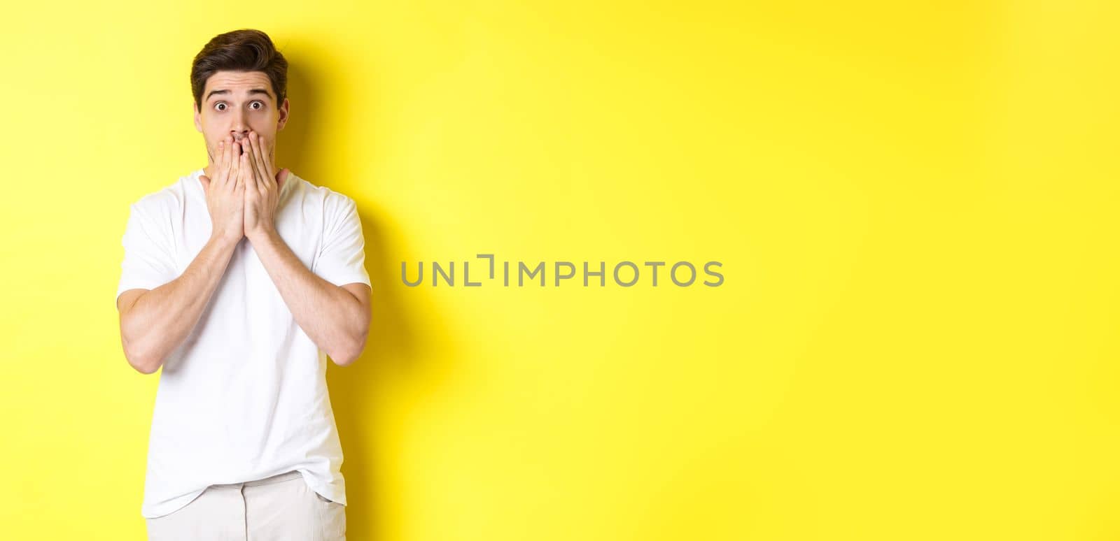 Handsome man looking shocked and speechless, holding hands on mouth, standing over yellow background.