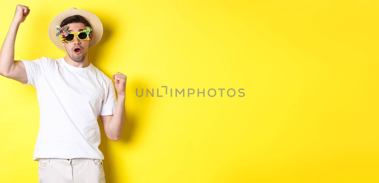 Concept of tourism and lifestyle. Happy guy tourist enjoying trip, rooting for you, fist pump and triumphing, going on journey in summer hat and sunglasses, yellow background.