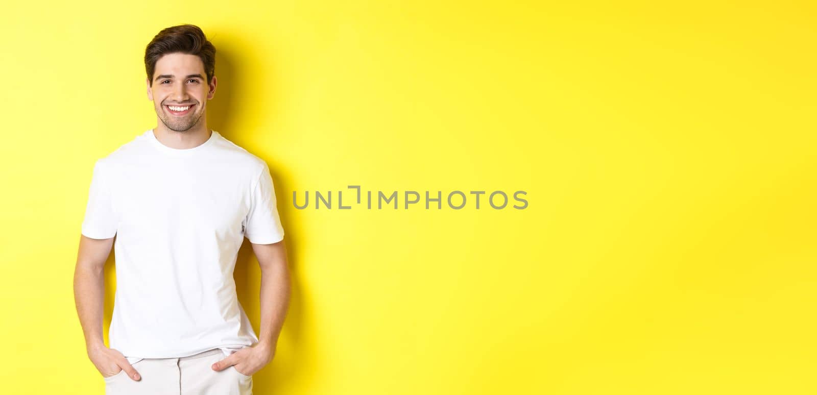 Young handsome man smiling at camera, holding hands in pockets, standing against yellow background.