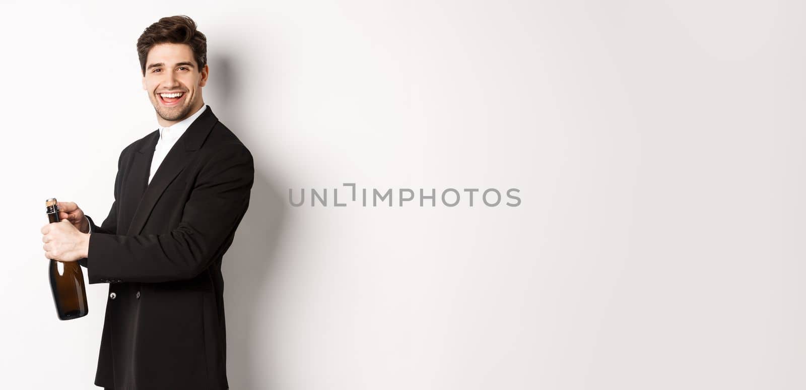 Handsome smiling man in trendy suit open a bottle of champagne, celebrating holidays, standing over white background.