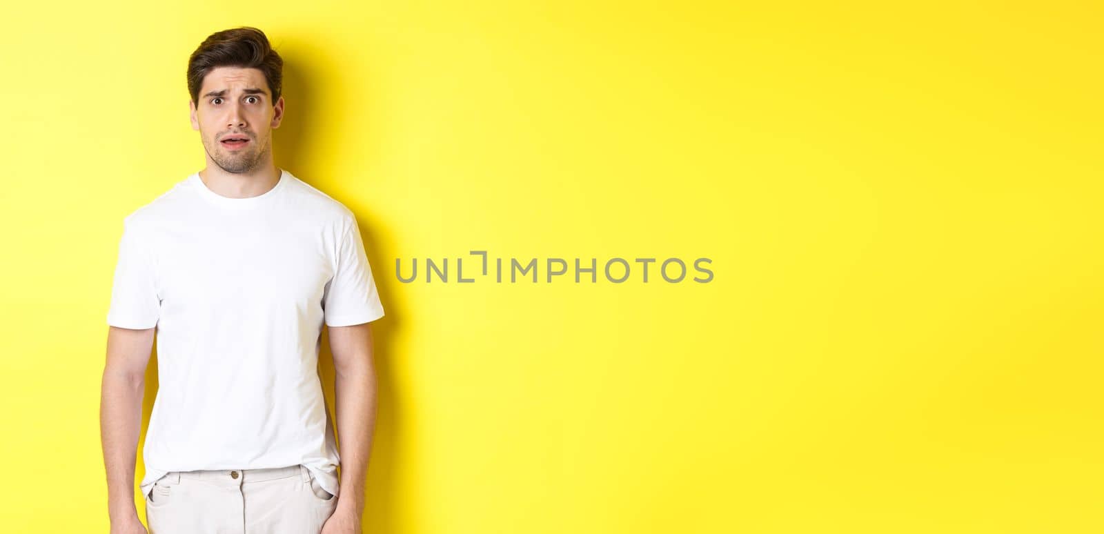 Image of confused and nervous man looking at something strange, frowning anxious, standing against yellow background.