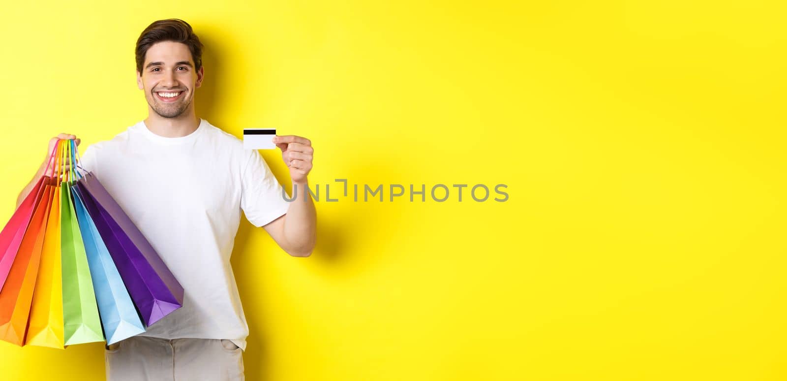 Young man shopping for holidays, holding paper bags and recommending bank credit card, standing over yellow background.