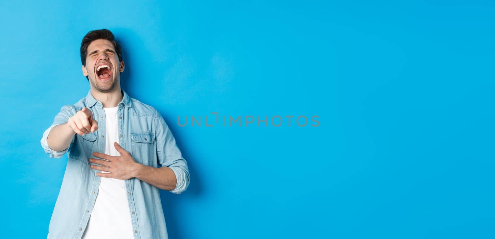 Adult man in casual outfit laughing out loud and pointing at you, looking at something funny, chuckling while standing against blue background.