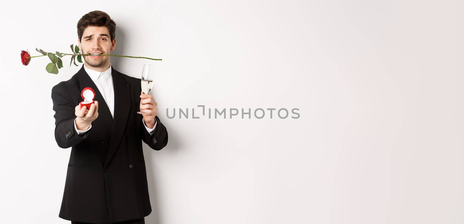 Romantic young man in suit making a proposal, holding rose in teeth and glass of champagne, showing engagement ring, asking to marry him, standing against white background.