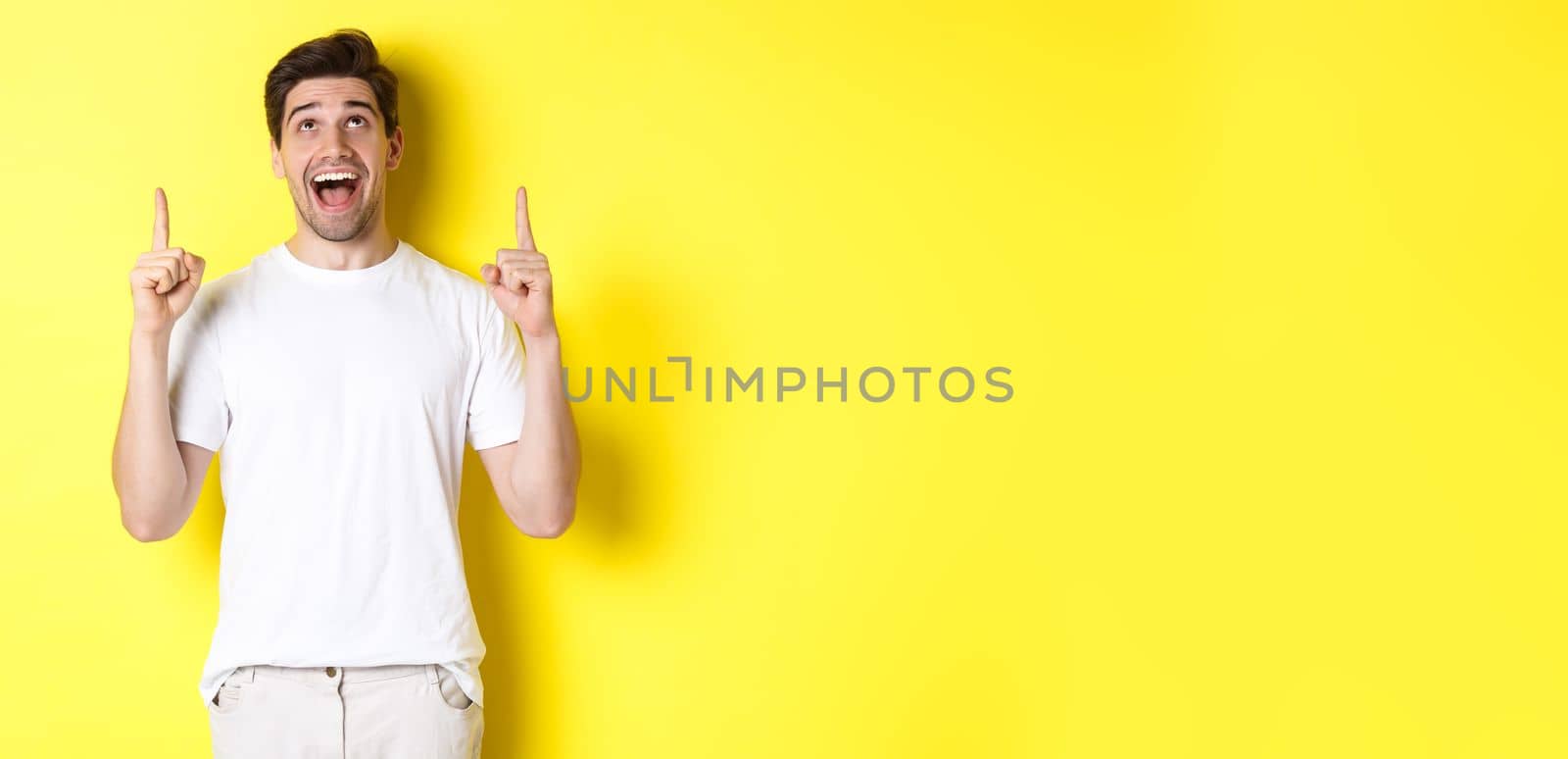 Cheerful young guy in white t-shirt reacting to promo offer, pointing and looking up with amazement, standing over yellow background.