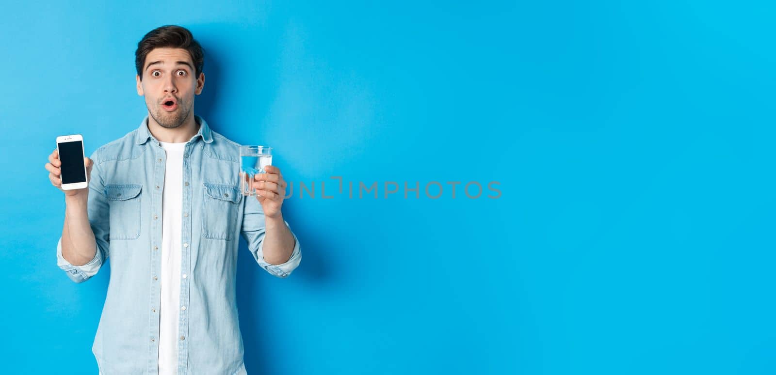 Man looking surprised, showing smartphone screen and glass of water, standing over blue background.