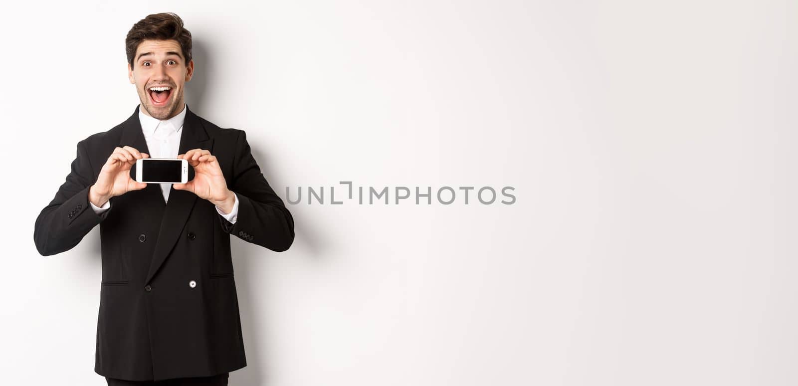 Image of cheerful, handsome man in black suit, showing smarthone screen and looking amazed, standing against white background.