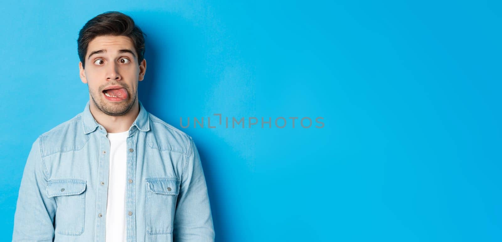 Close-up of young man making funny expressions, showing tongue and looking at camera, standing over blue background. Copy space