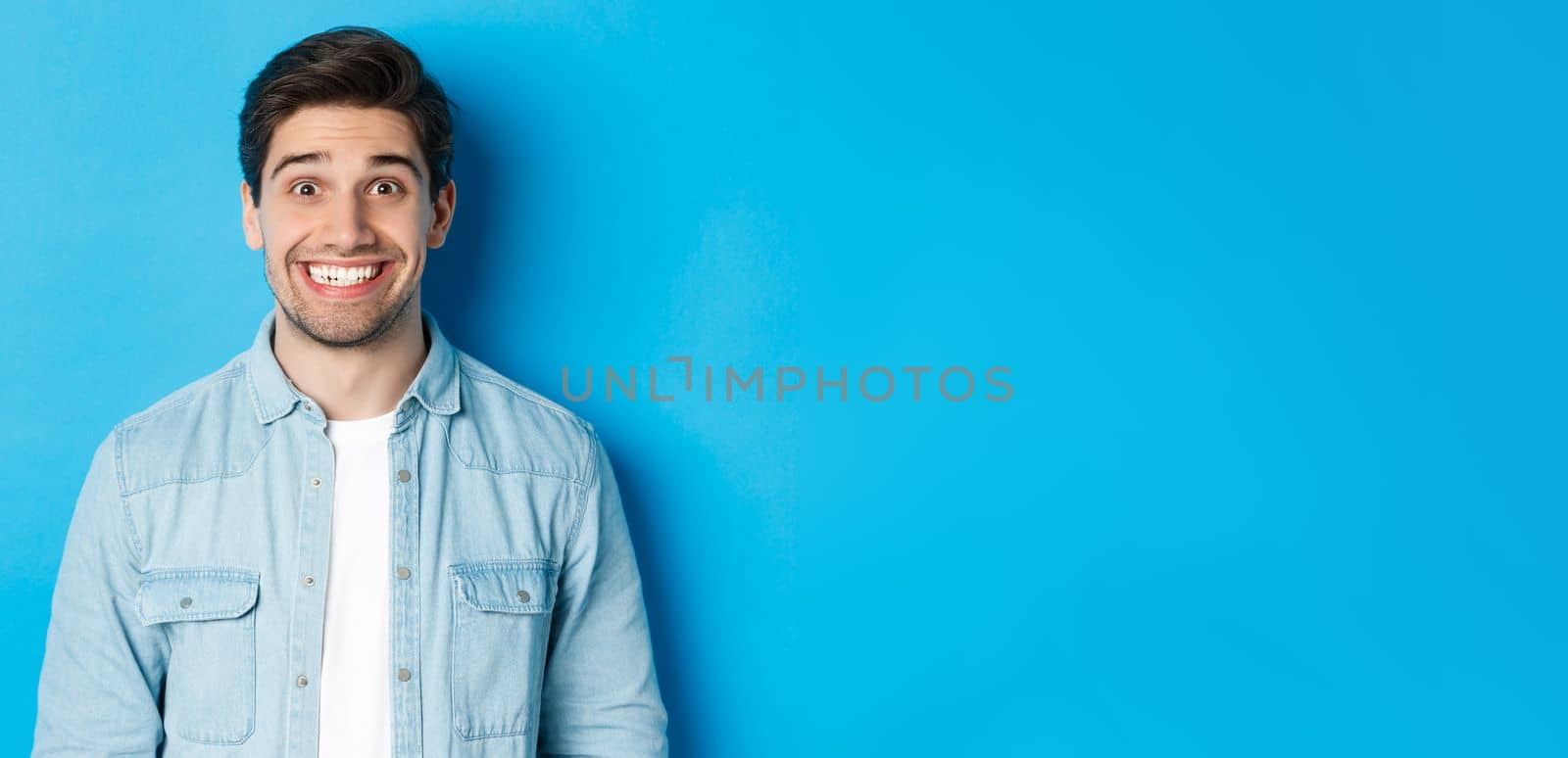 Close-up of smiling excited man with beard, looking amused at advertisement, standing against blue background by Benzoix