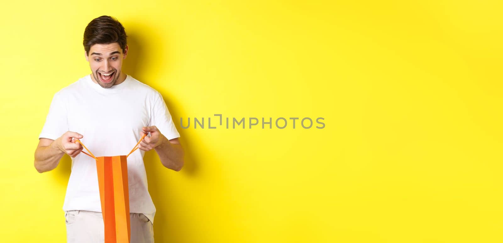 Excited guy open bag with gift, looking inside with amazement and happy face, standing against yellow background.