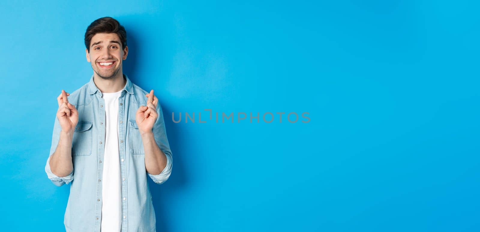 Handsome hopeful man making a wish, crossing fingers and smiling, waiting for results, standing against blue background.