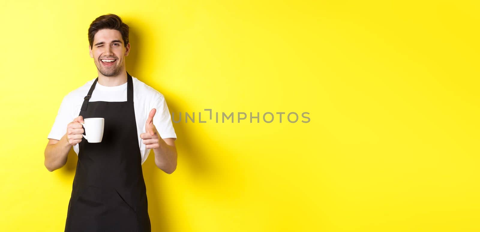 Handsome barista in black apron holding coffee cup, pointing finger at you, inviting visit cafe, standing over yellow background.