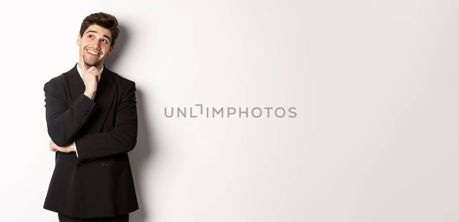 Portrait of thoughtful smiling man in stylish party suit, looking at upper left corner and thinking, imaging things, standing over white background.