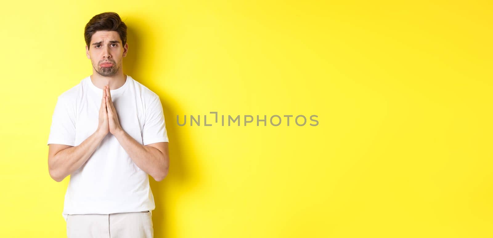 Sad guy begging for something, sulking and asking for favour, saying sorry, standing over yellow background.