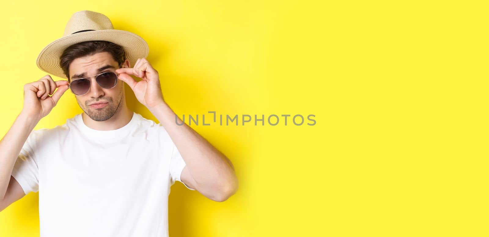 Concept of tourism and vacation. Close-up of cool tourist enjoying holidays on trip, wearing sunglasses with straw hat, yellow background.