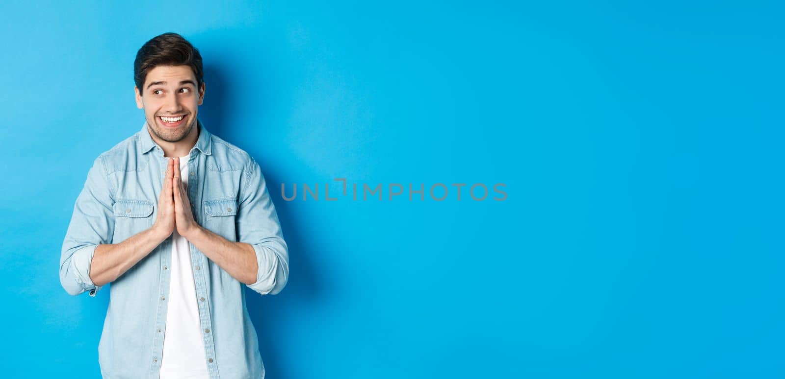 Hopeful smiling man looking left, holding hands in pray, begging for something or pleading, standing against blue background.