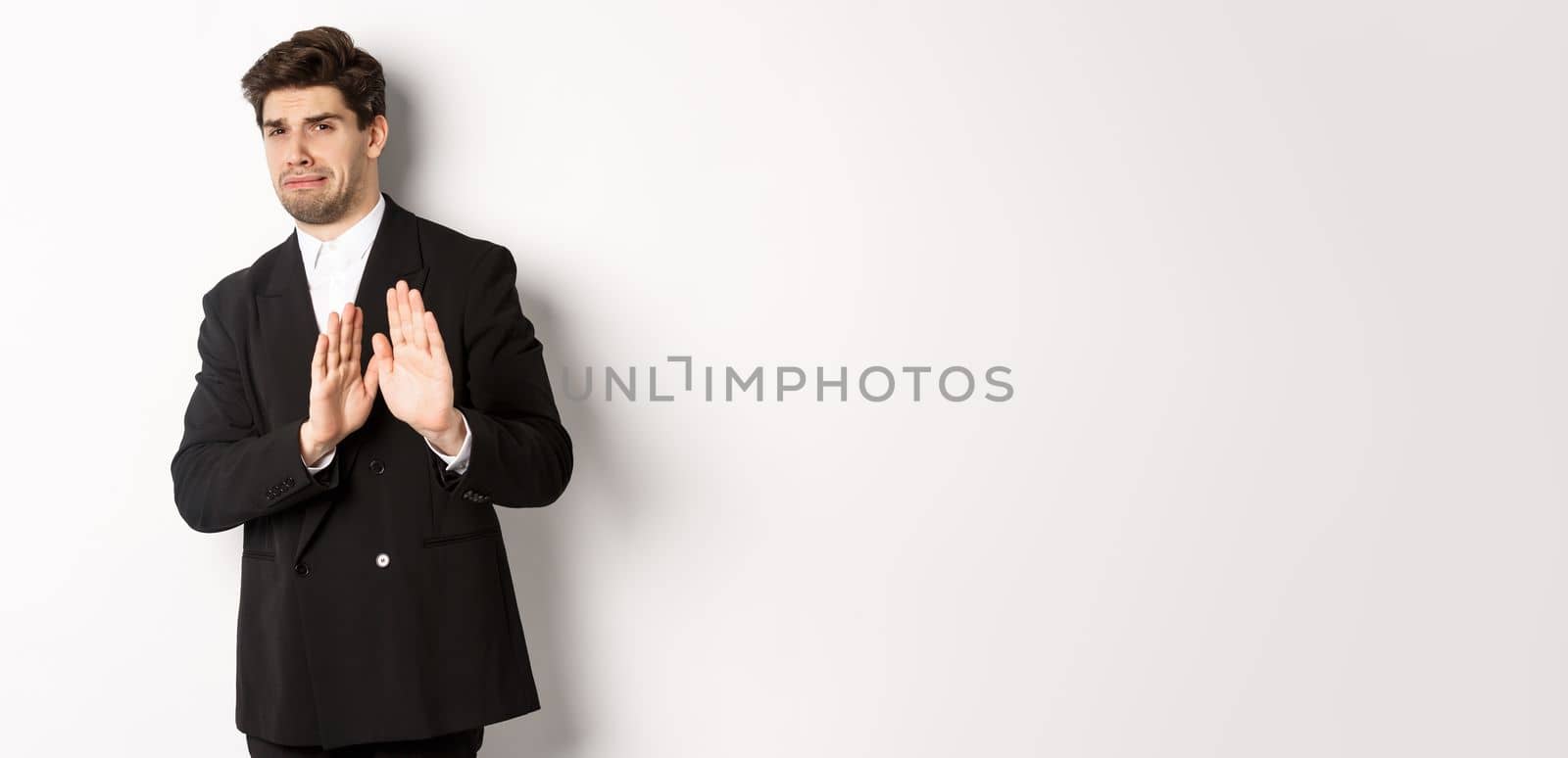 Image of stylish handsome man in black suit, cringe from something embarrassing, declining offer and showing stop gesture, standing against white background.