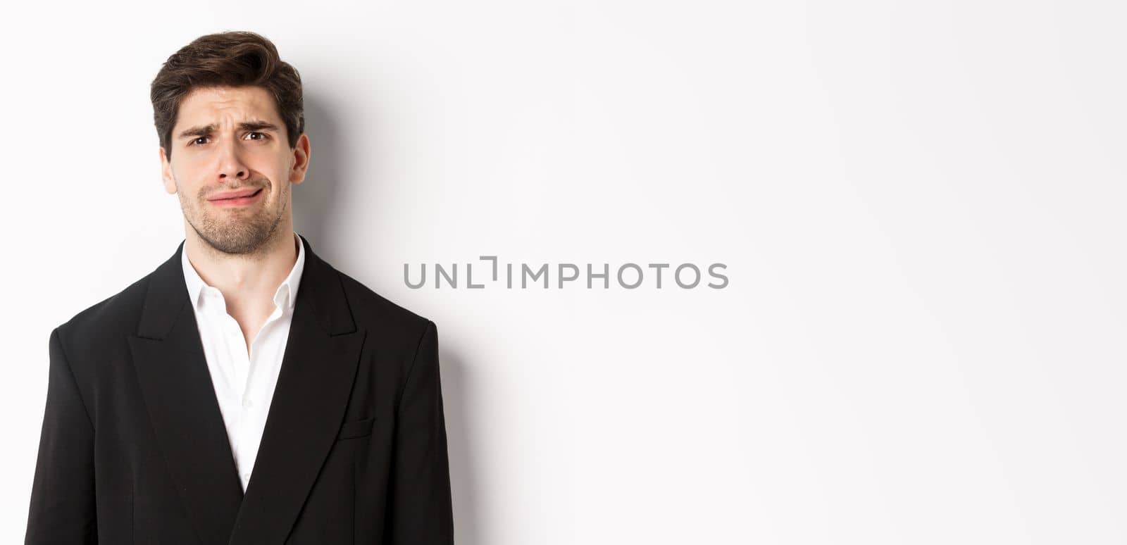 Close-up of confused handsome man in suit, grimacing and looking perplexed, wearing trendy suit, standing against white background.