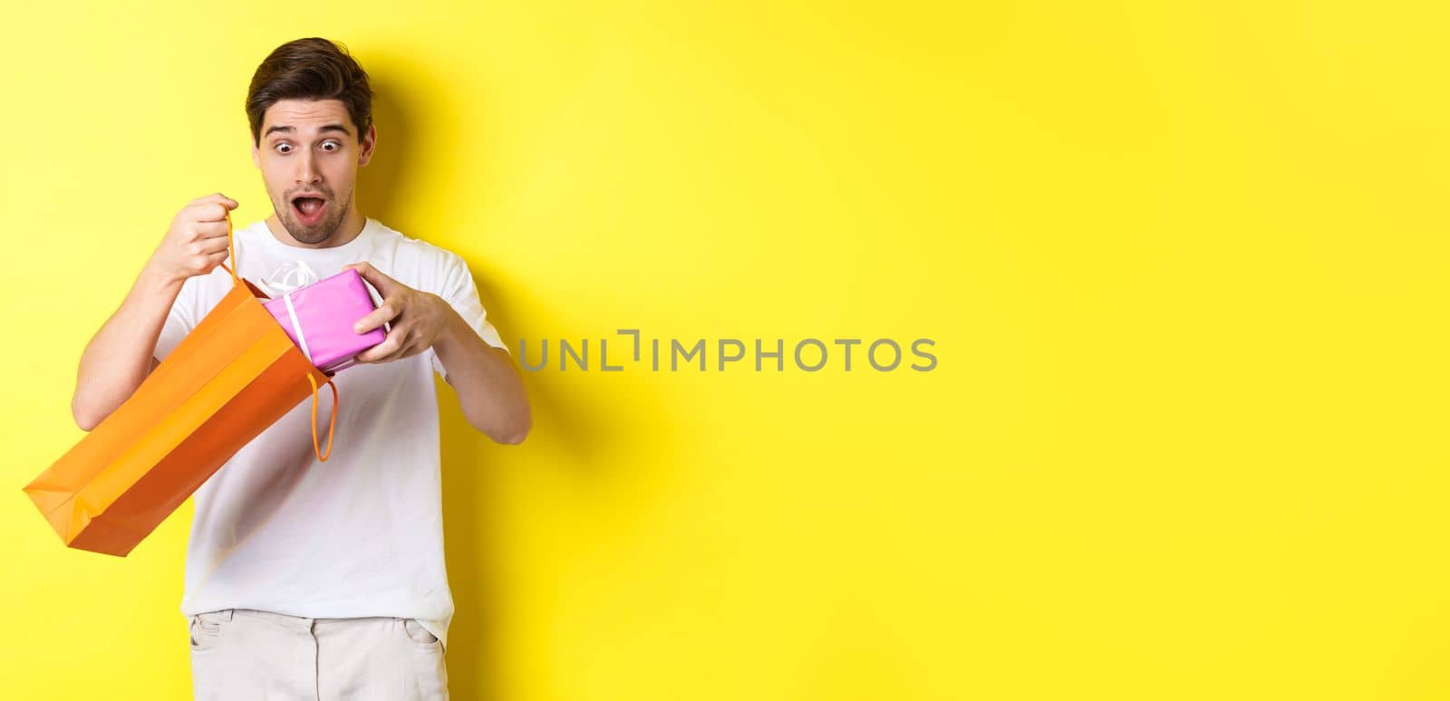 Concept of holidays and celebration. Young man looking surprised as take out gift from shopping bag, standing over yellow background.