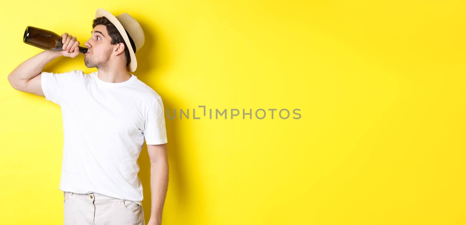 Concept of tourism and vacation. Man drinking wine from bottle on holidays, standing against yellow background by Benzoix