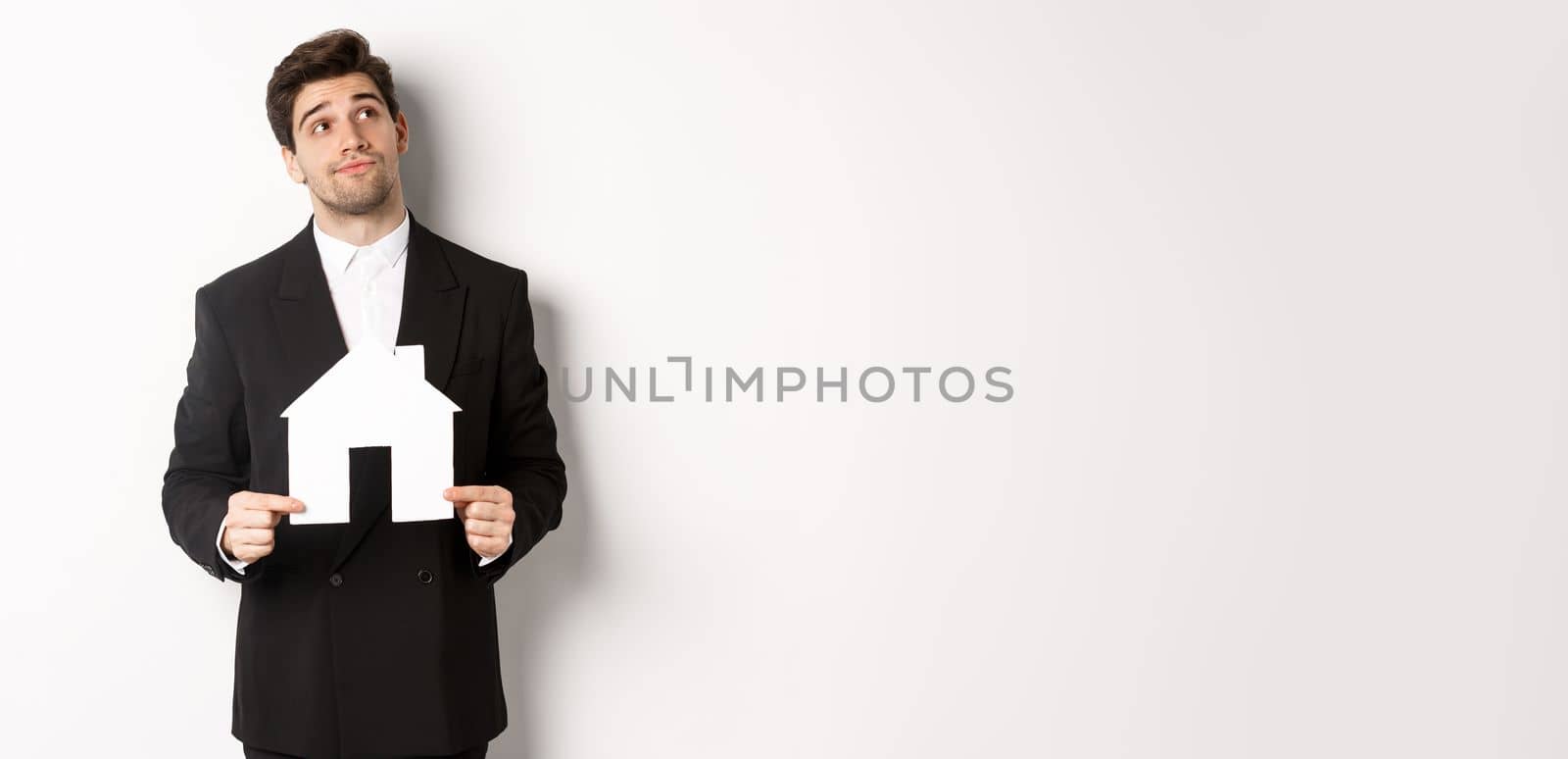 Image of handsome businessman in black suit, looking for home, holding house maket and gazing dreamy at upper right corner, standing against white background.