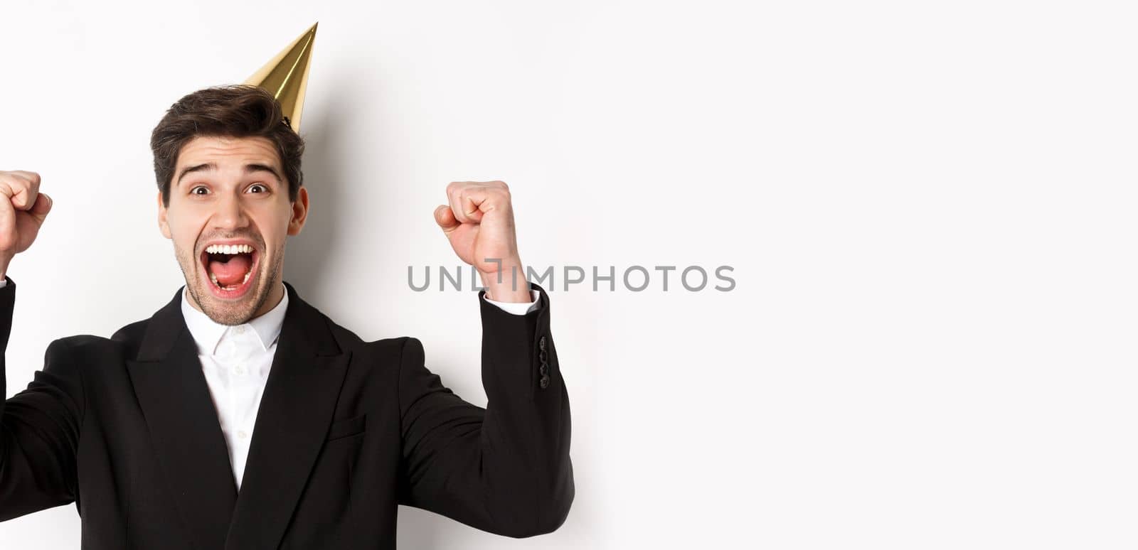 Close-up of happy good-looking man, wearing party hat and suit, raising hands up and rejoicing, celebrating new year, standing against white background.