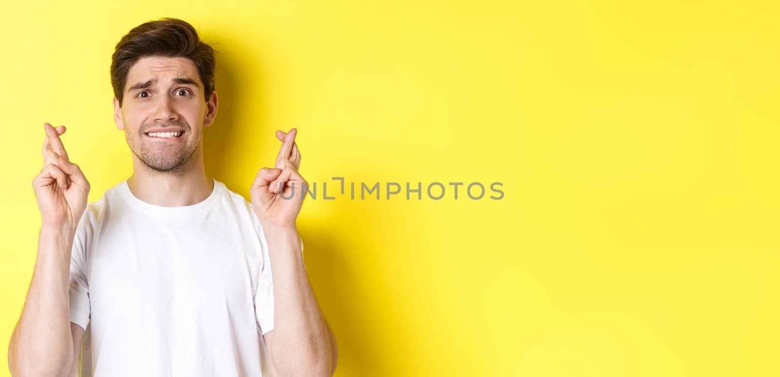 Close-up of nervous man making wish, holding fingers crossed and biting lip worried, standing over yellow background.